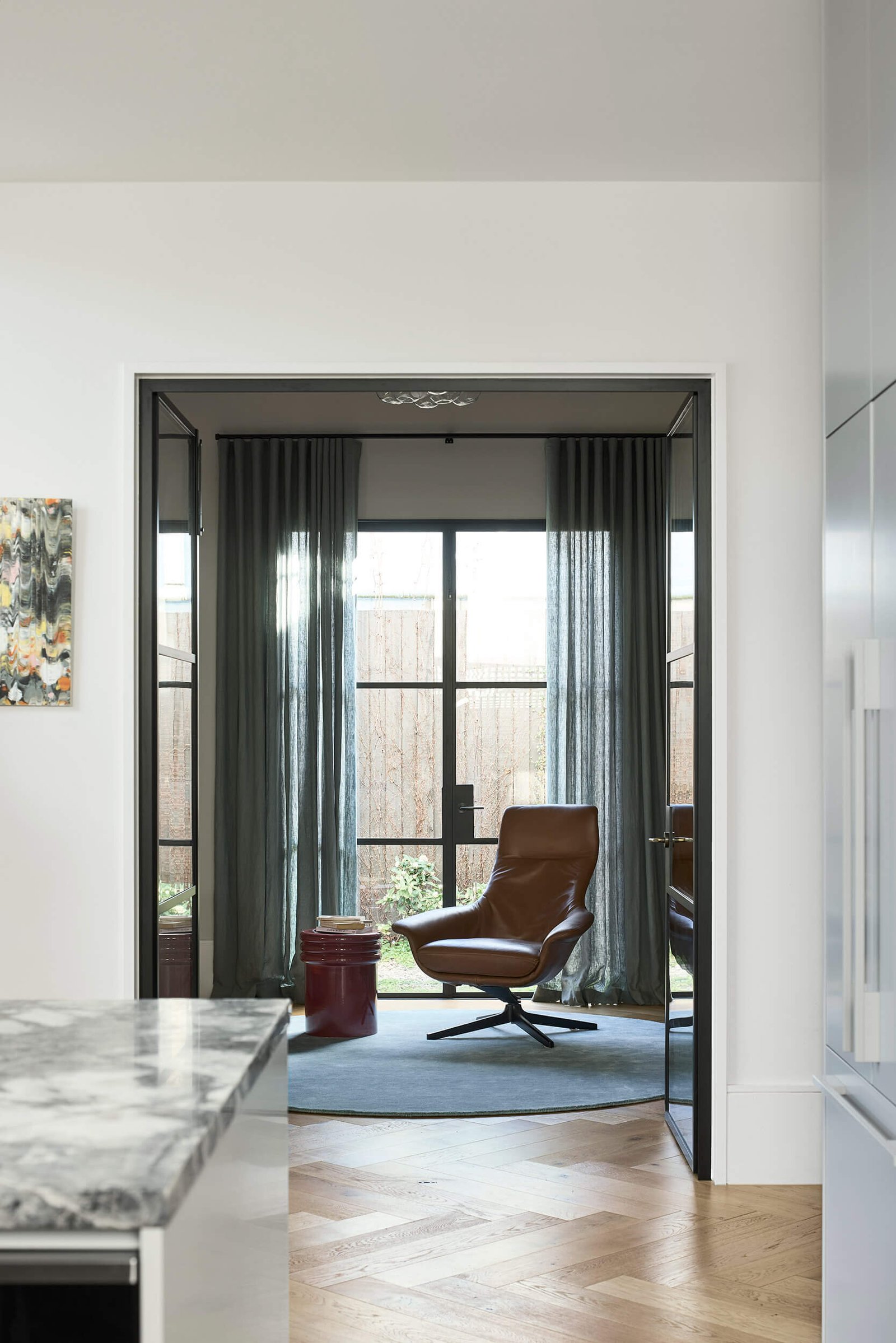 A glass-framed doorway leads into a small yet refined home office where sheer curtains diffuse the brightness, balancing privacy with openness.