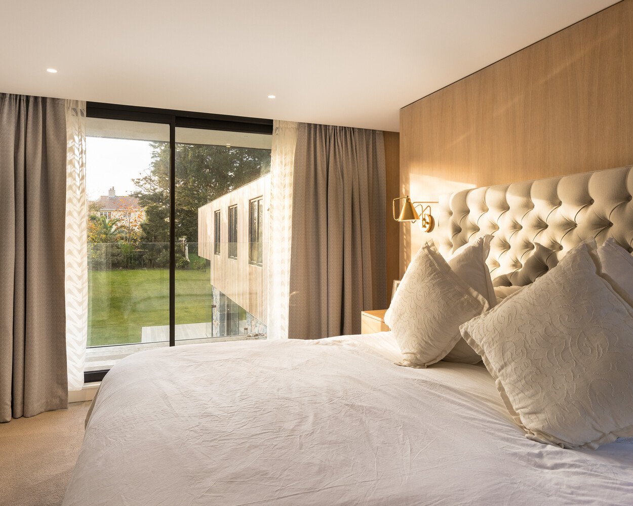 At the top of the stairs, the hallway leads to a tranquil master suite. A wood accent wall sits behind the bed, adding warmth to the otherwise pared-back space. Through a sliding glass door, a small balcony opens to views of the surrounding garden, a quiet reminder that nature is always near.