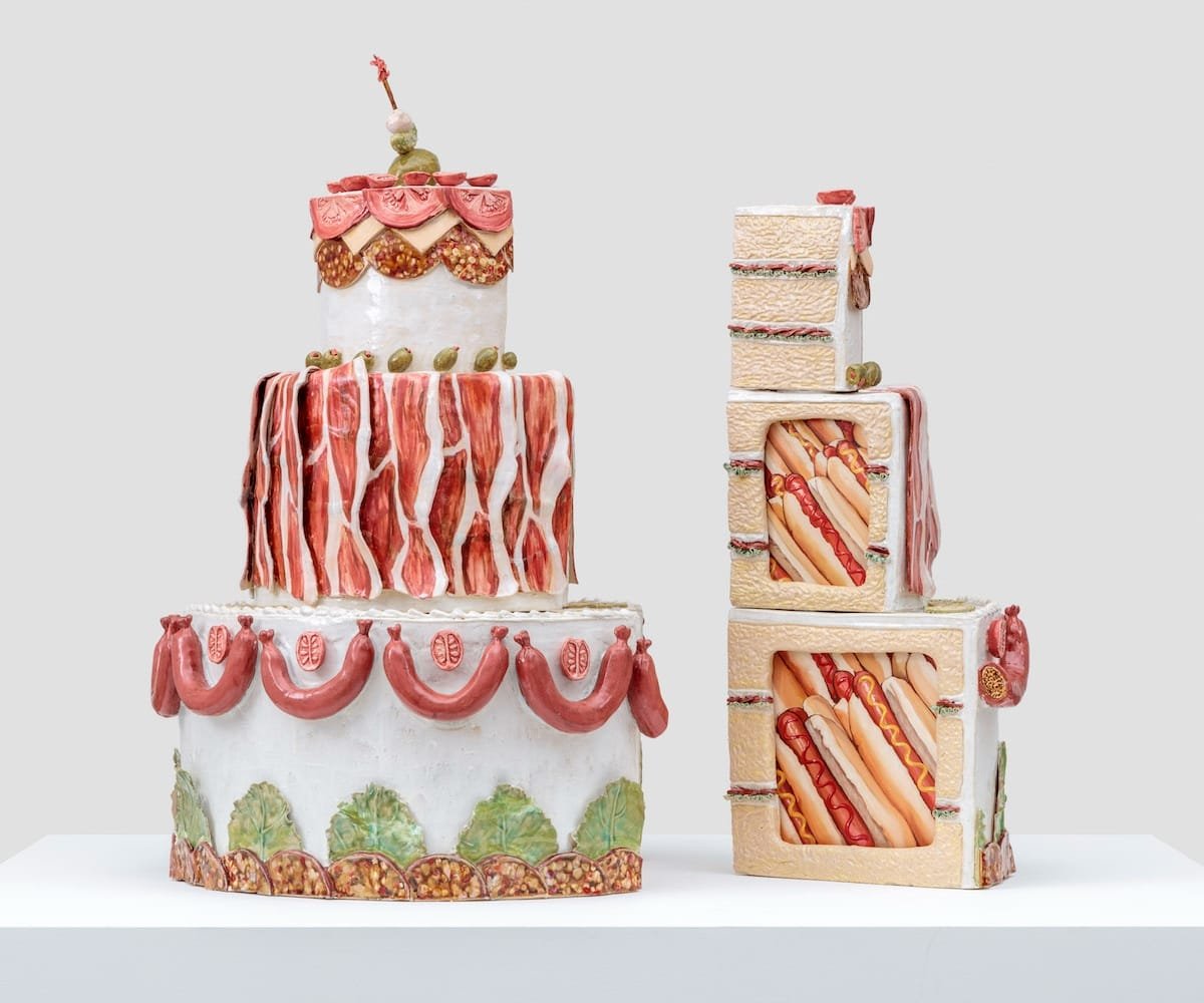 two ceramic cake sculptures by Stephanie Temma Heir with bacon, sausages, hot dogs, and other meats embedded in the sides
