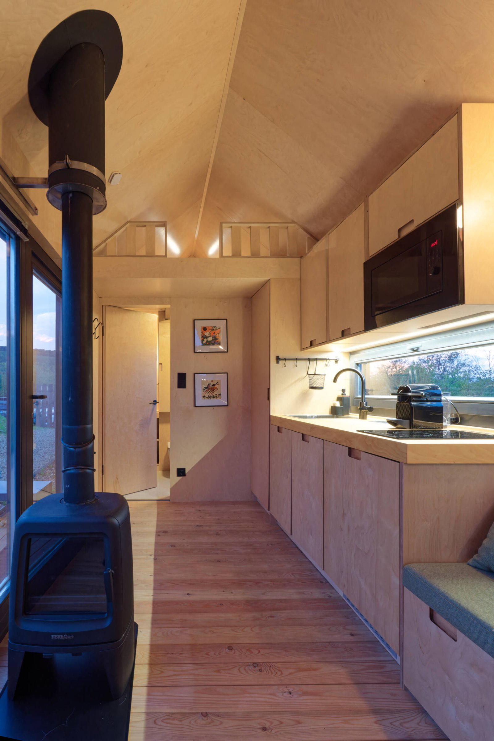 Light birch plywood, a visible gabled roof, and a striking trapezoidal window make this micro cabin interior feel surprisingly spacious. Included in the cabin is a sleeping area, lounge, kitchen, loft, and bathroom.
