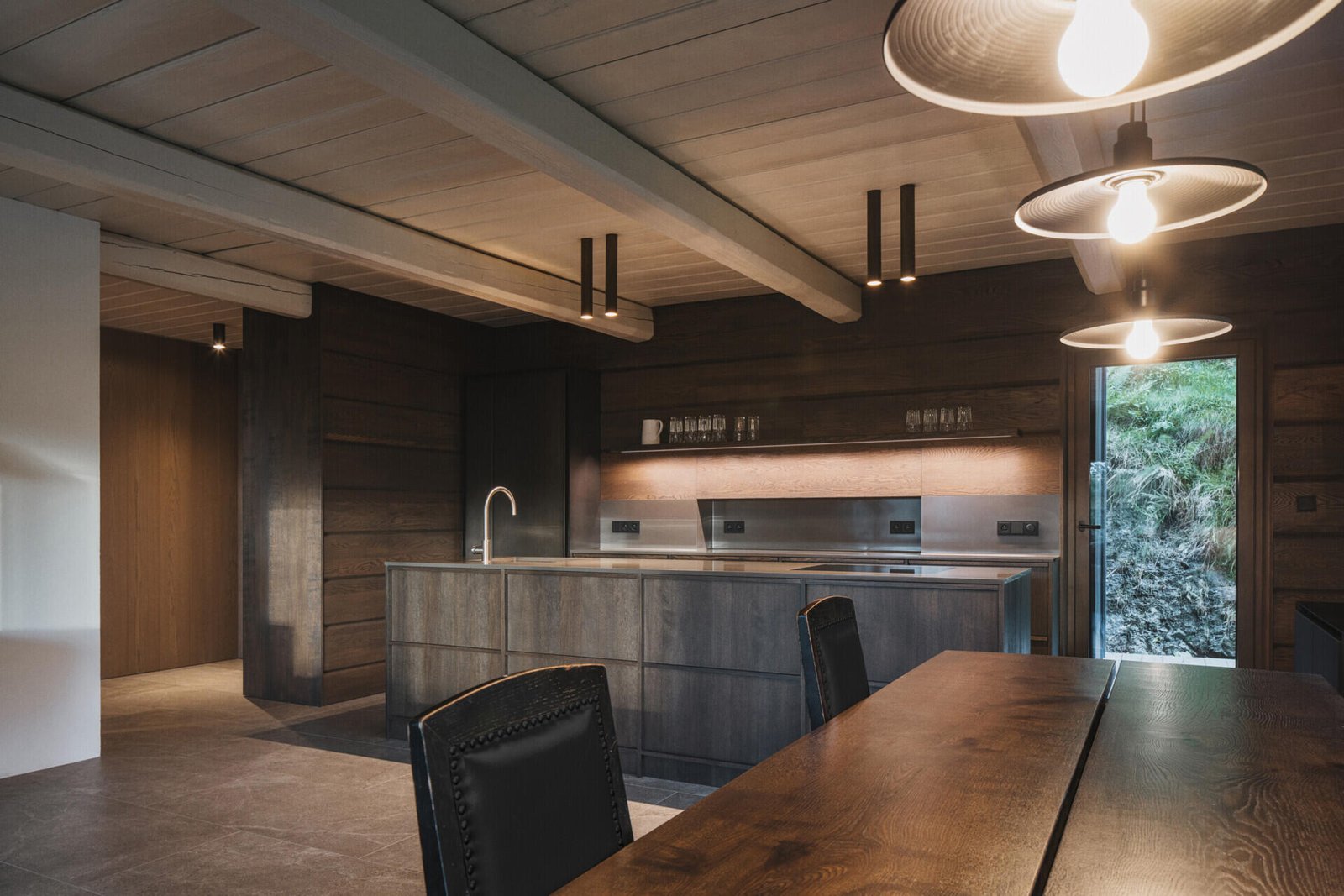At the heart of this mountain cottage, the main living room retains the scale dictated by an original beam ceiling. Built-in seating runs along the windows, and a large table anchors the space for meals and gatherings. Nearby, the kitchen with its dark cabinetry, almost blends into the background.