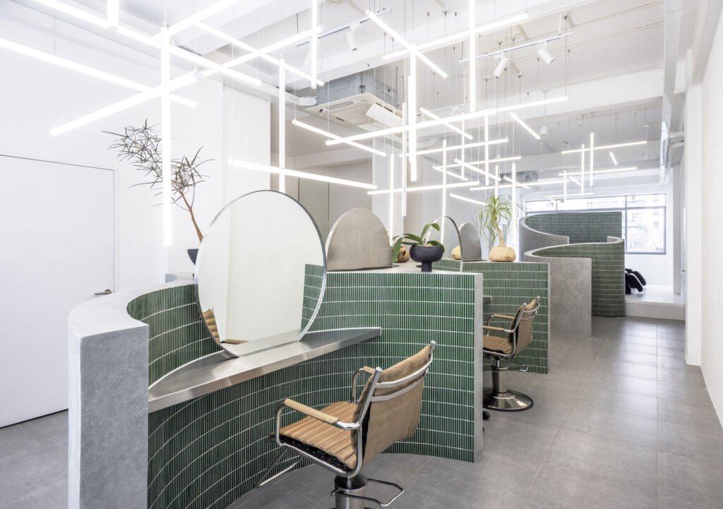 A hair salon in Ho Chi Minh City designed by Inrestudio, shaped by a continuous sculptural wall that guides movement through waiting areas, styling stations, and shampoo spaces. The interior shifts between green tile and raw grey surfaces, with light and form changing as you move through each level of the maisonette.
