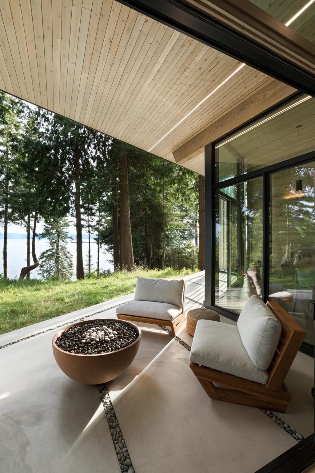 A minimalist living room with a gray sofa and black fireplace opens through sliding glass doors to a covered patio with water views.