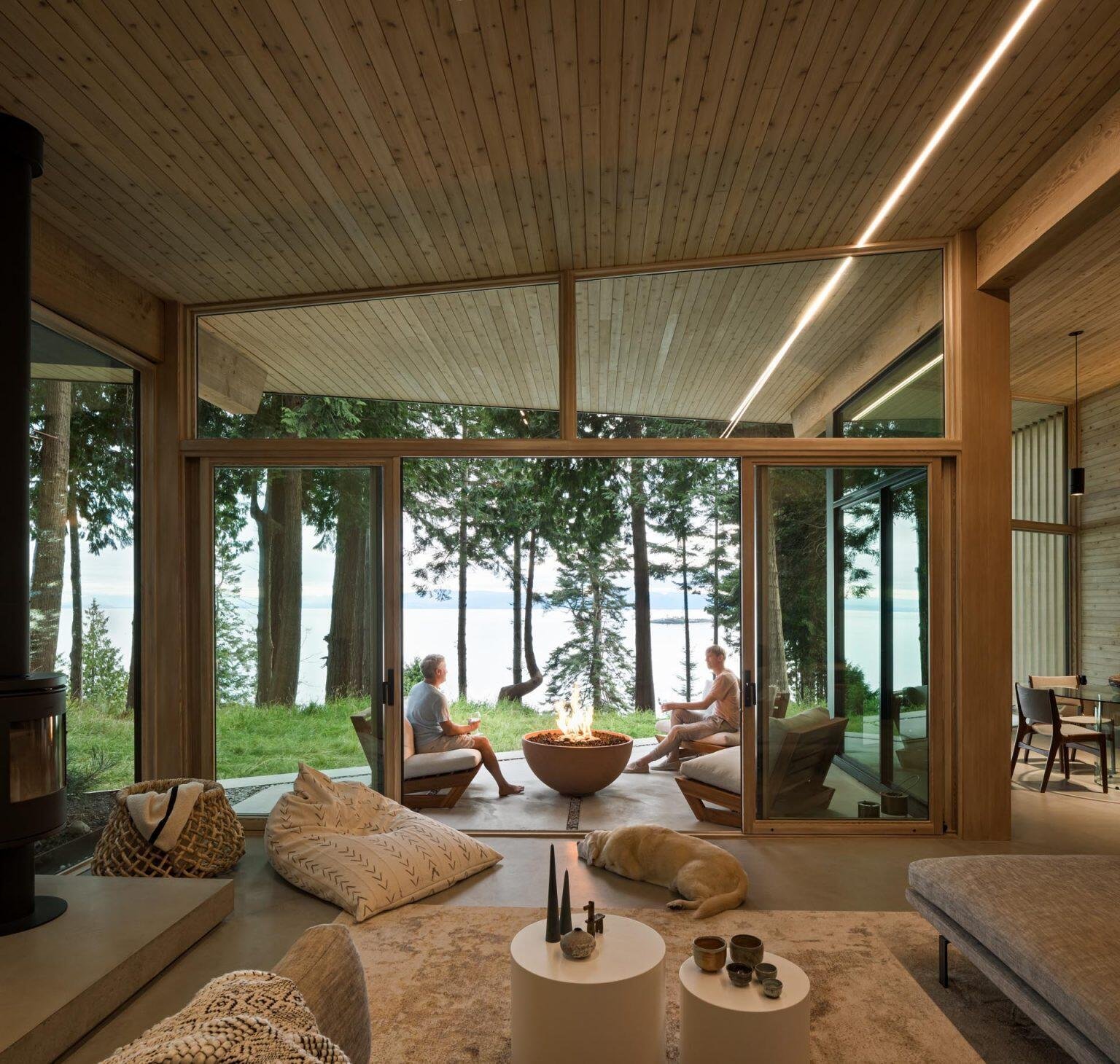 A minimalist living room with a gray sofa and black fireplace opens through sliding glass doors to a covered patio with water views.