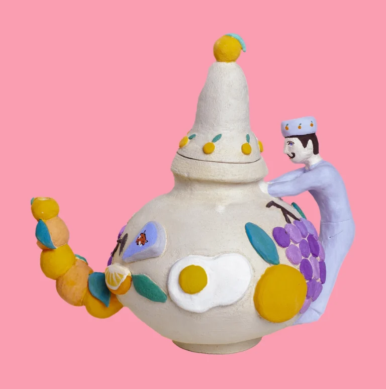 Whimsical ceramic teapot decorated with colourful fruits and a small figurine, set against a pink background